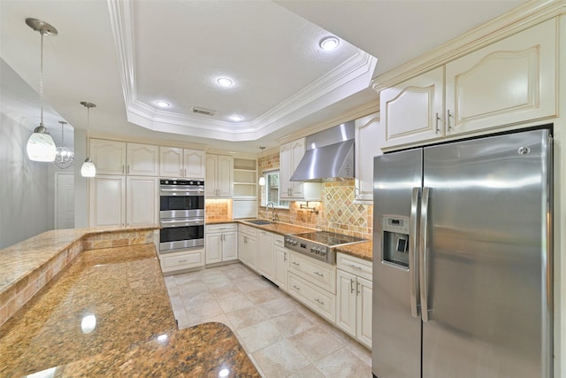 kitchen with appliances with stainless steel finishes, a raised ceiling, wall chimney exhaust hood, and cream cabinetry