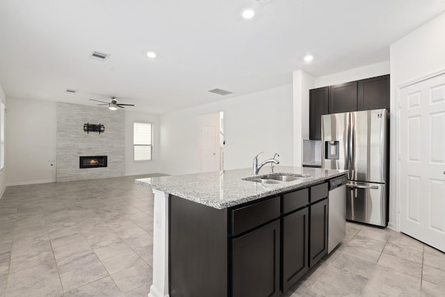 kitchen featuring appliances with stainless steel finishes, light stone countertops, a kitchen island with sink, a fireplace, and a sink