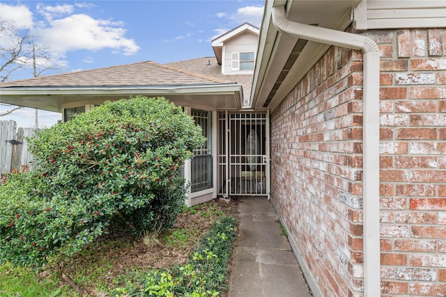 doorway to property featuring brick siding, roof with shingles, and fence