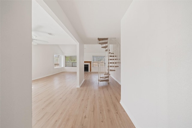 unfurnished living room featuring ceiling fan, a fireplace, baseboards, stairway, and light wood-type flooring