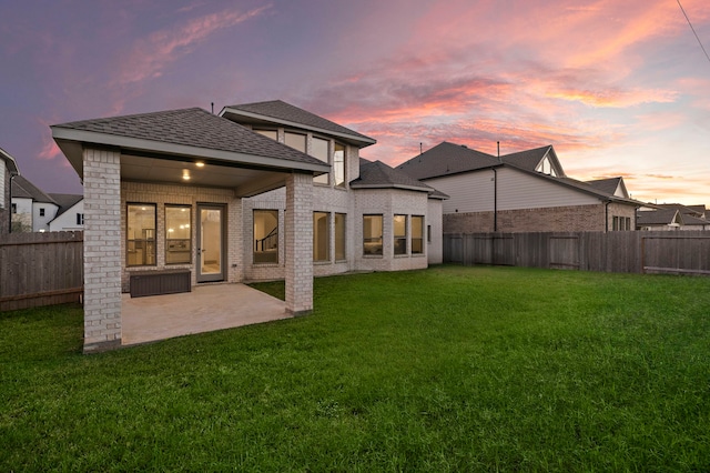 back of property at dusk with a shingled roof, a fenced backyard, a yard, a patio area, and brick siding