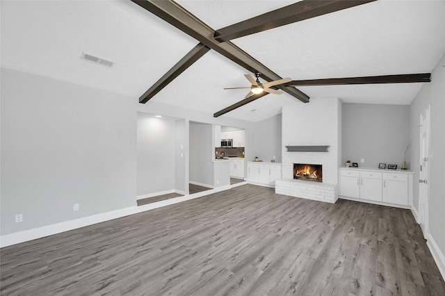 unfurnished living room with visible vents, lofted ceiling with beams, a brick fireplace, wood finished floors, and baseboards