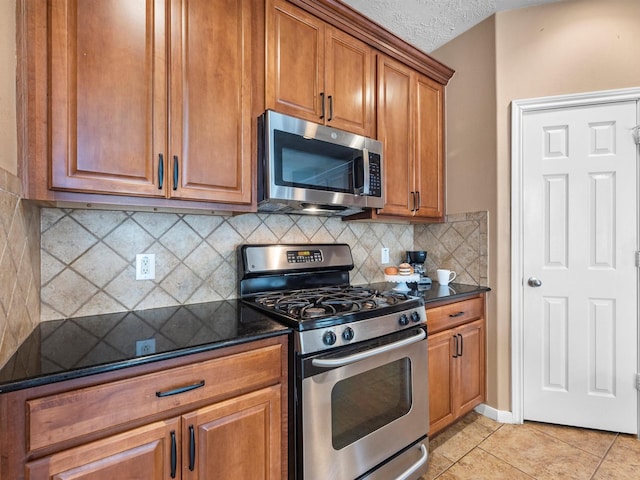 kitchen featuring light tile patterned floors, brown cabinetry, decorative backsplash, appliances with stainless steel finishes, and dark stone countertops