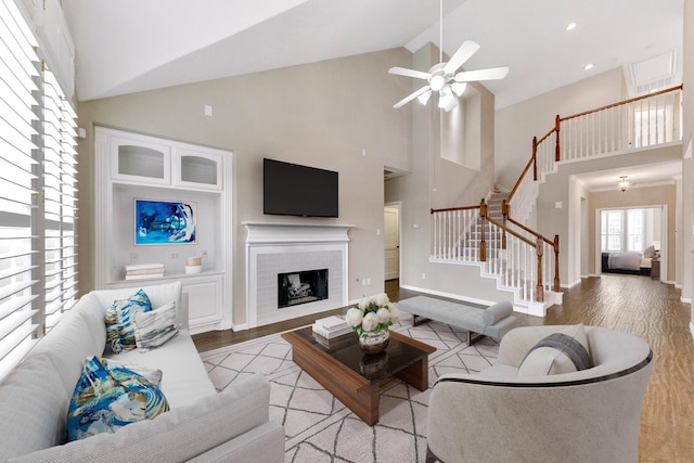 living area with high vaulted ceiling, light wood-style flooring, a fireplace, a ceiling fan, and stairs