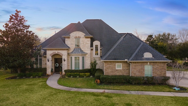 french country style house with stone siding, a front lawn, roof with shingles, and brick siding