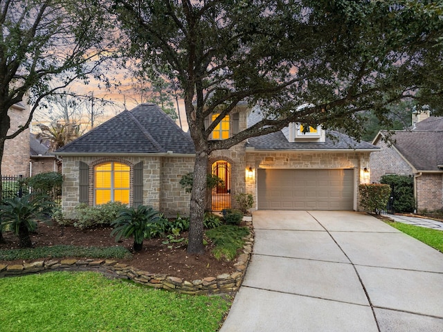 french country inspired facade with a shingled roof, concrete driveway, stone siding, and an attached garage