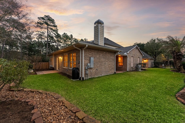 back of property at dusk with a yard, brick siding, a chimney, and fence