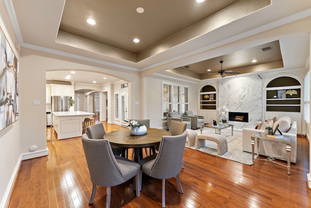 dining room with ceiling fan, a fireplace, light wood-style flooring, and a tray ceiling