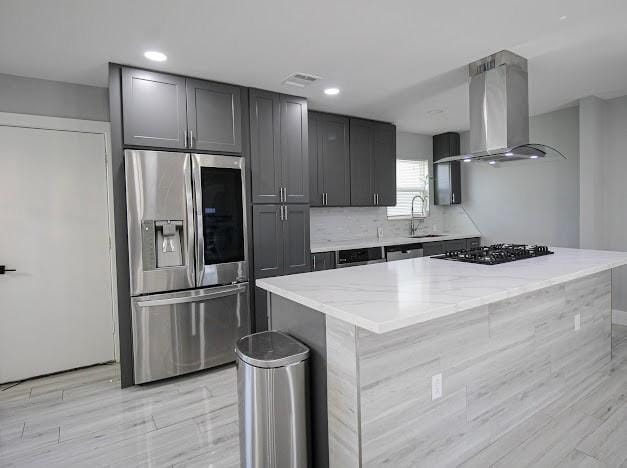 kitchen with wall chimney range hood, light stone counters, stainless steel refrigerator with ice dispenser, and a sink