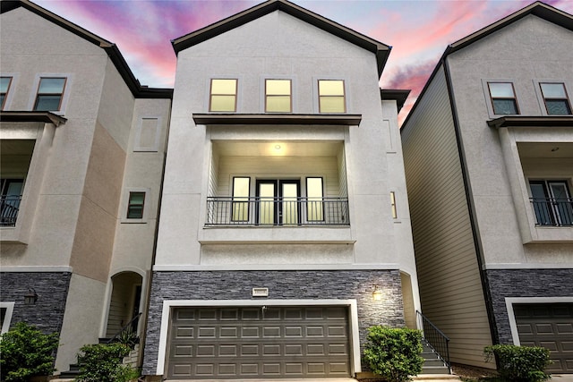 view of front of house with driveway, a balcony, stone siding, an attached garage, and stucco siding