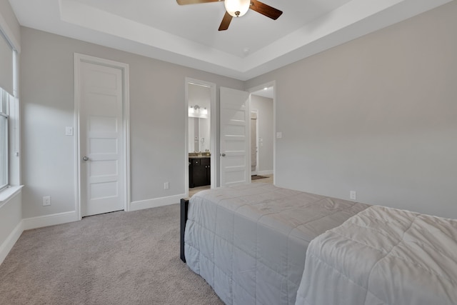 carpeted bedroom featuring a ceiling fan, a tray ceiling, ensuite bath, and baseboards