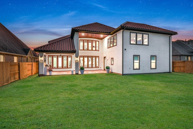 back of house at dusk with a tiled roof, a fenced backyard, a lawn, and french doors