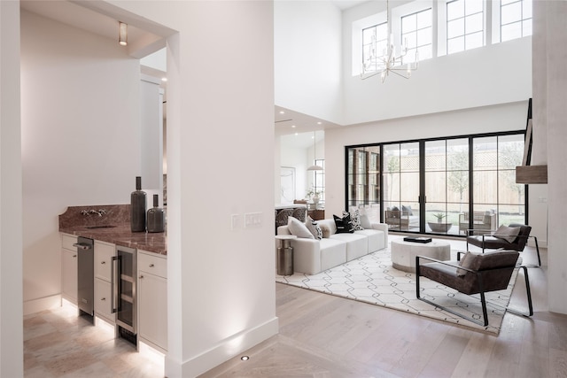 living room with wine cooler, a towering ceiling, light wood-style flooring, an inviting chandelier, and a bar