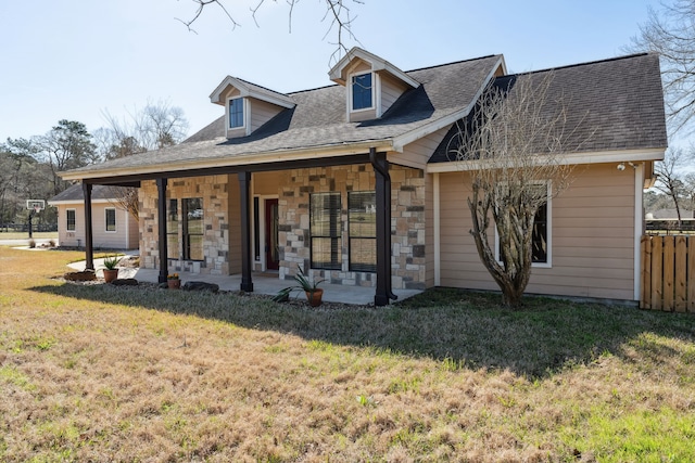 view of front facade with a patio, stone siding, roof with shingles, fence, and a front yard