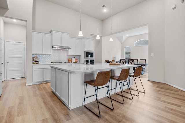 kitchen featuring light wood finished floors, tasteful backsplash, a towering ceiling, white cabinets, and under cabinet range hood