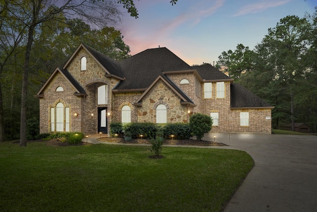 french country inspired facade with concrete driveway, brick siding, a lawn, and a shingled roof