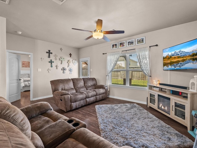 living area featuring dark wood-type flooring, a glass covered fireplace, baseboards, and a ceiling fan