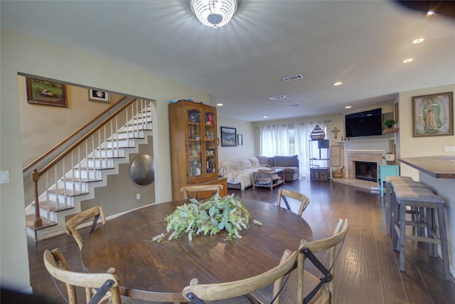 dining room with a fireplace with raised hearth, recessed lighting, visible vents, stairs, and dark wood finished floors