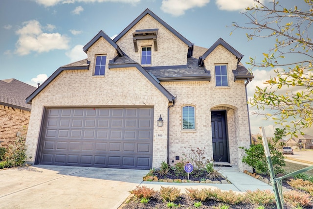 french country inspired facade featuring driveway, an attached garage, a shingled roof, and brick siding