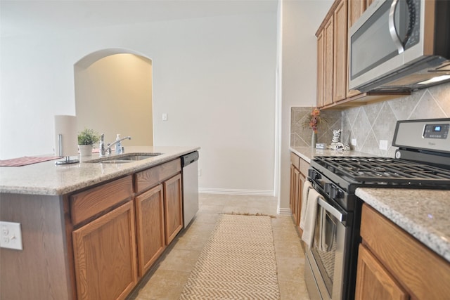 kitchen with light stone counters, stainless steel appliances, decorative backsplash, brown cabinetry, and a sink