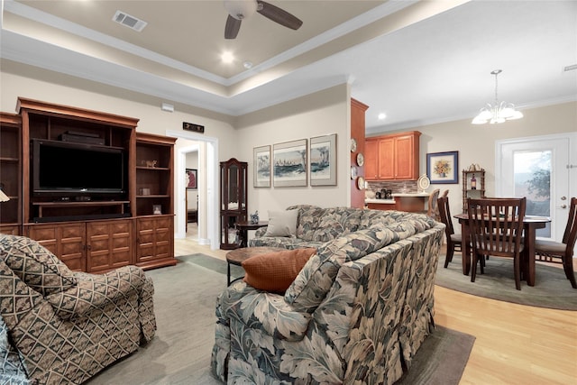 living area with ceiling fan with notable chandelier, visible vents, light wood-style floors, ornamental molding, and a tray ceiling