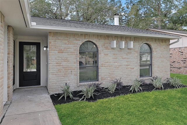 doorway to property featuring brick siding, a yard, and roof with shingles