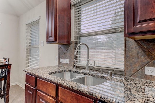 kitchen with reddish brown cabinets, tasteful backsplash, a sink, dark stone countertops, and baseboards