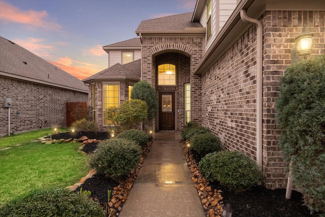 property entrance with roof with shingles, brick siding, and a lawn