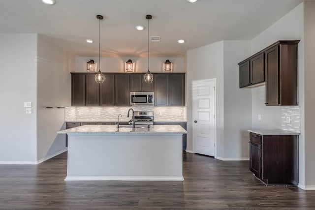 kitchen featuring dark brown cabinetry, dark wood finished floors, appliances with stainless steel finishes, light stone countertops, and a kitchen island with sink