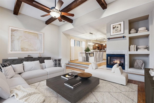 living area with built in shelves, beam ceiling, a tiled fireplace, ceiling fan, and wood finished floors