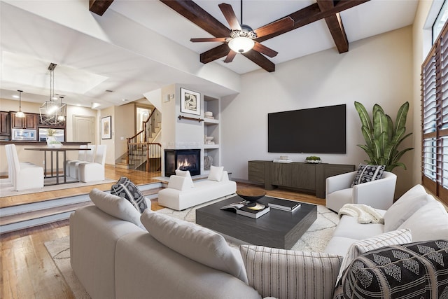 living area featuring built in shelves, a glass covered fireplace, light wood-type flooring, beamed ceiling, and stairs