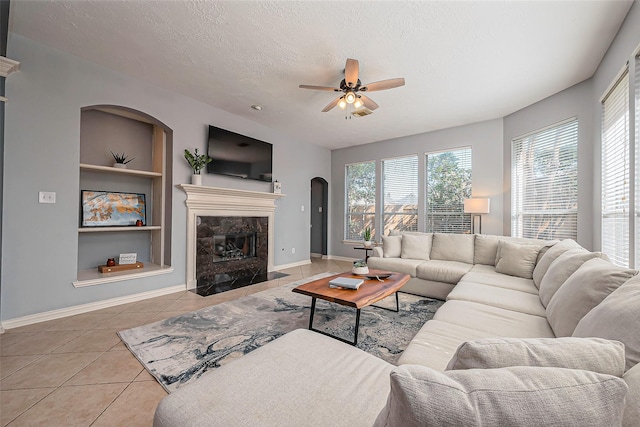 tiled living room featuring built in features, a high end fireplace, ceiling fan, a textured ceiling, and baseboards