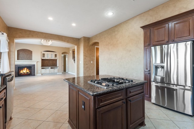 kitchen with light tile patterned floors, appliances with stainless steel finishes, a kitchen island, dark stone counters, and a lit fireplace