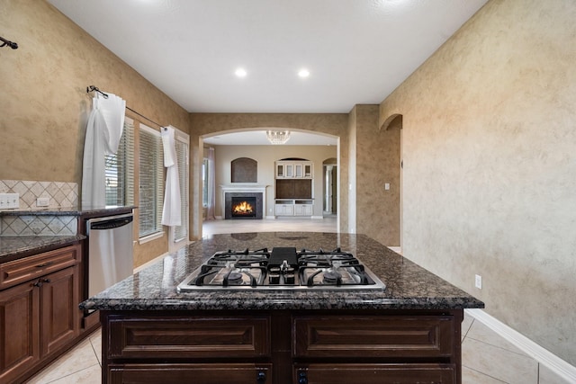 kitchen featuring light tile patterned floors, appliances with stainless steel finishes, dark stone countertops, a lit fireplace, and backsplash