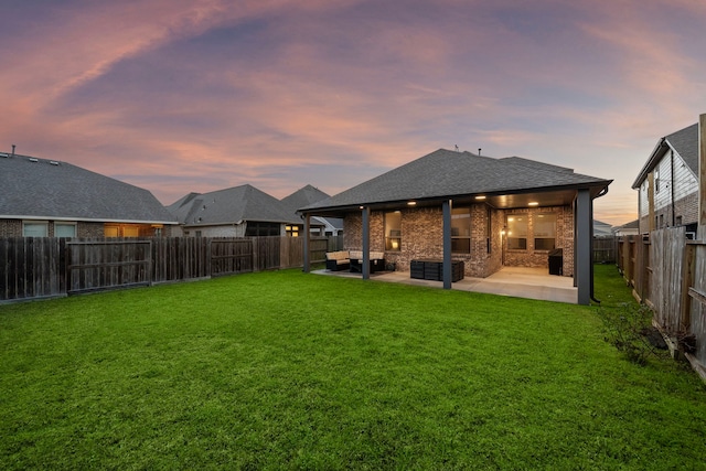 back of house at dusk with a patio, a fenced backyard, a shingled roof, a lawn, and brick siding