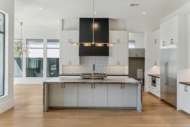 kitchen with built in refrigerator, visible vents, white cabinets, light wood-type flooring, and backsplash