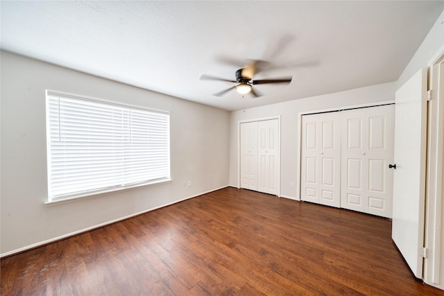 unfurnished bedroom featuring dark wood-style floors, ceiling fan, baseboards, and two closets