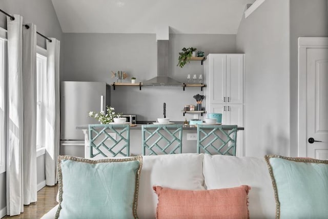 kitchen featuring lofted ceiling, freestanding refrigerator, wall chimney range hood, white cabinetry, and open shelves