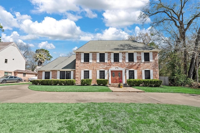 colonial-style house with driveway, roof with shingles, a front lawn, and brick siding