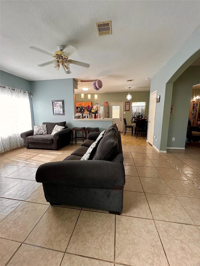 tiled living room with arched walkways, a textured ceiling, ceiling fan with notable chandelier, visible vents, and baseboards