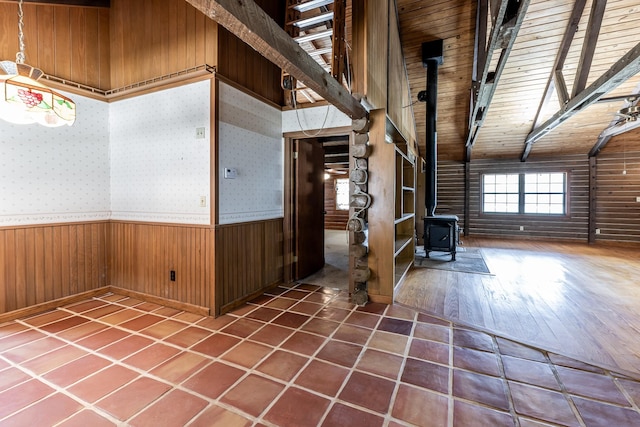 unfurnished room featuring wallpapered walls, a wainscoted wall, beamed ceiling, a wood stove, and wood walls