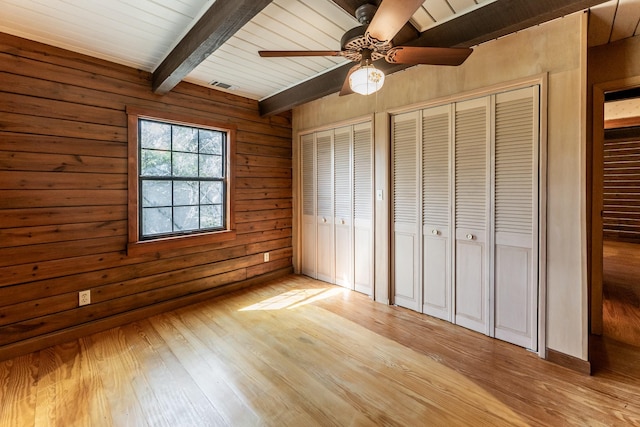 unfurnished bedroom with wooden walls, visible vents, light wood-style flooring, beamed ceiling, and multiple closets