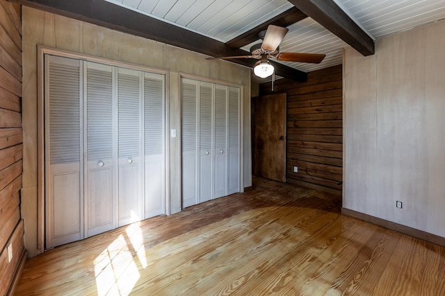 unfurnished bedroom featuring light wood finished floors, wooden walls, a ceiling fan, two closets, and beam ceiling