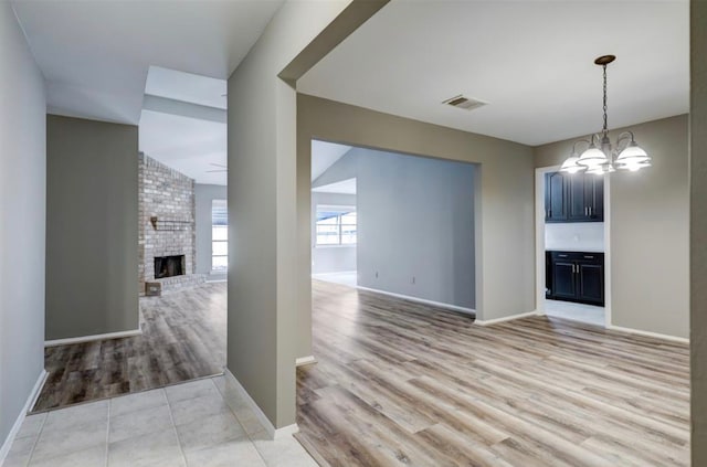 unfurnished living room with light wood finished floors, visible vents, a brick fireplace, and an inviting chandelier