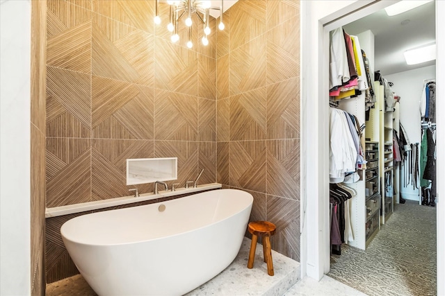 bathroom featuring a walk in closet, a soaking tub, a notable chandelier, and tile walls