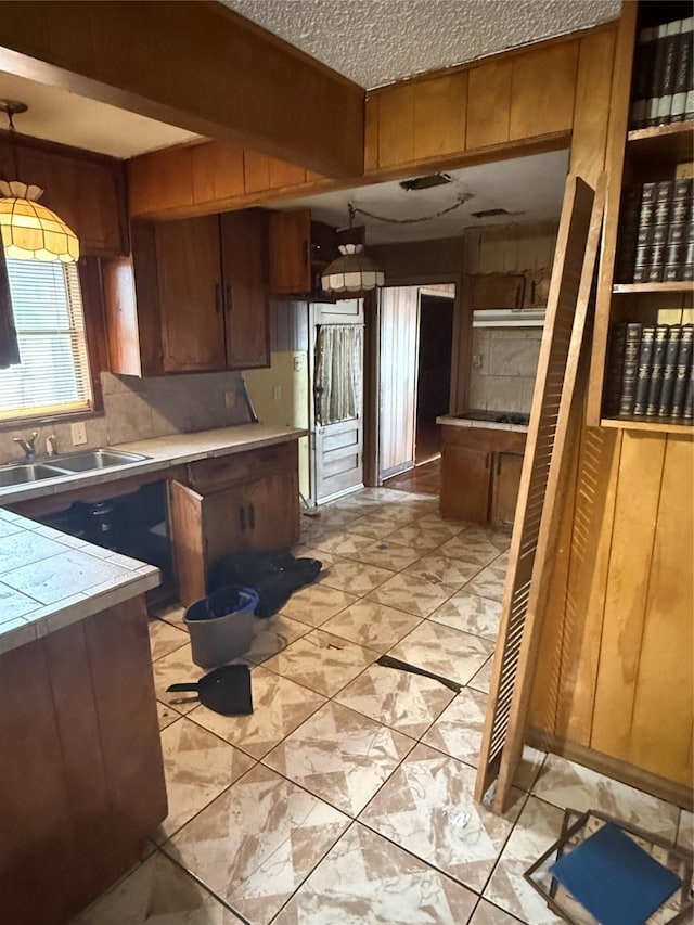 kitchen featuring brown cabinetry, tile countertops, a sink, a textured ceiling, and backsplash