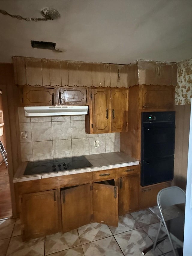 kitchen with tile countertops, tasteful backsplash, brown cabinetry, under cabinet range hood, and black appliances