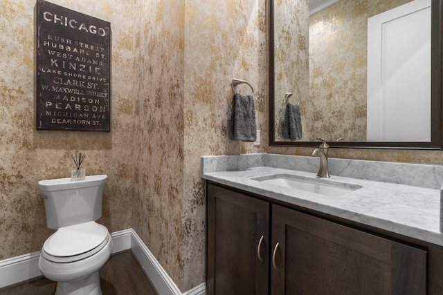 half bath with toilet, baseboards, vanity, and wallpapered walls