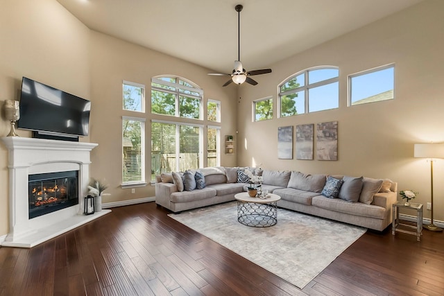 living room with baseboards, a ceiling fan, a glass covered fireplace, dark wood-style floors, and a high ceiling