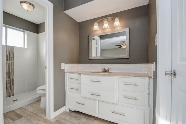 bathroom with toilet, a ceiling fan, a tile shower, vanity, and baseboards